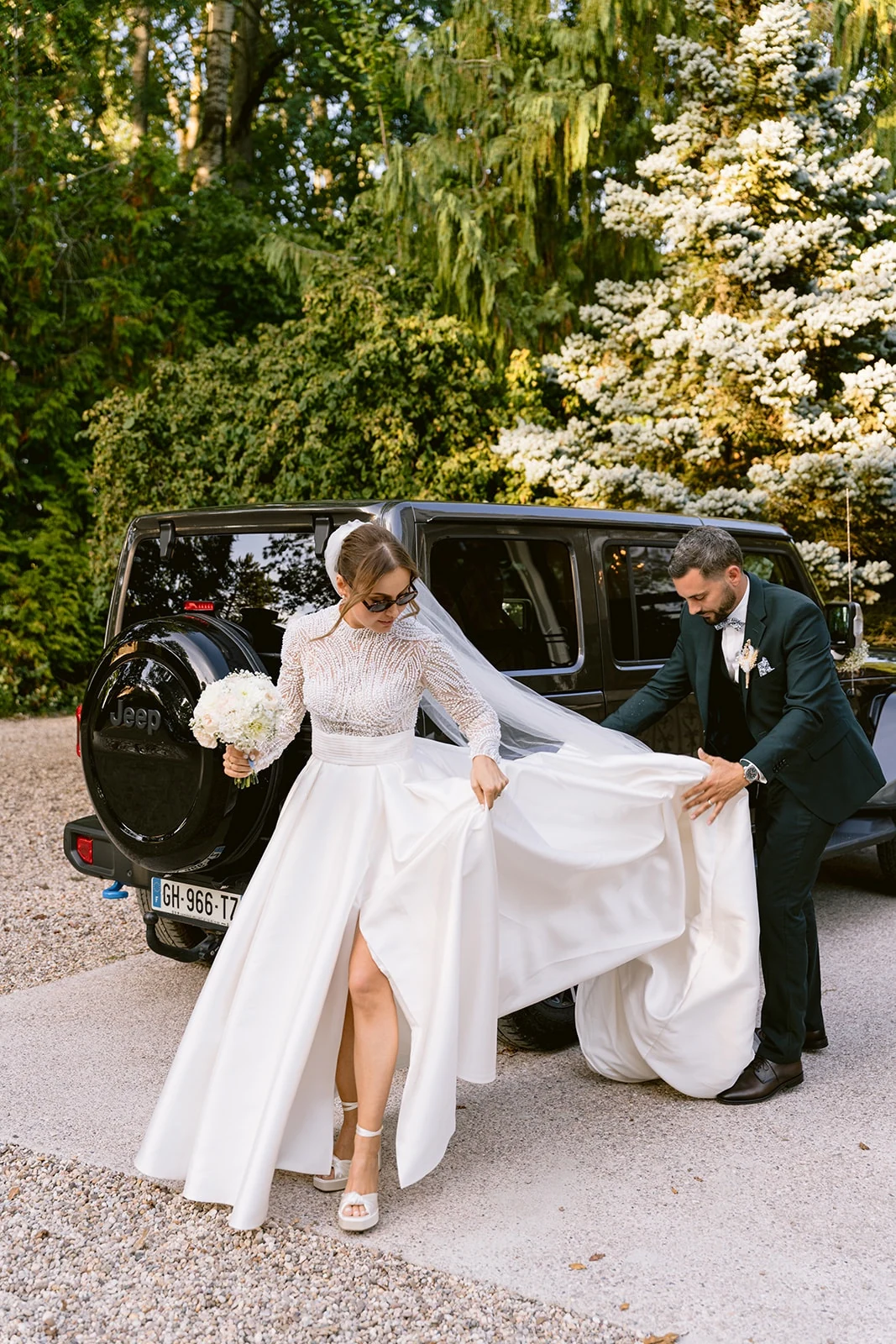 Le marié soulève la traîne de la mariée près d’un véhicule noir, dans un jardin.