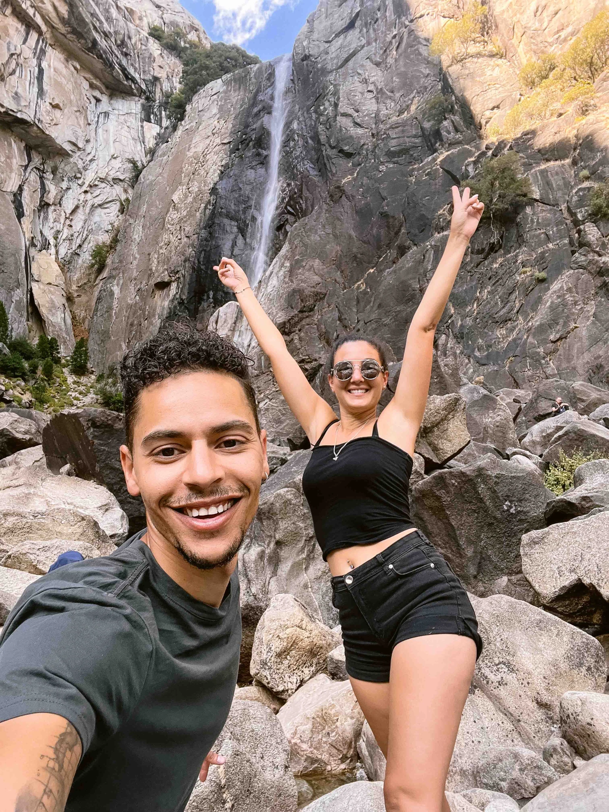 Un couple souriant se photographie au pied d’une cascade, la femme levant les bras.