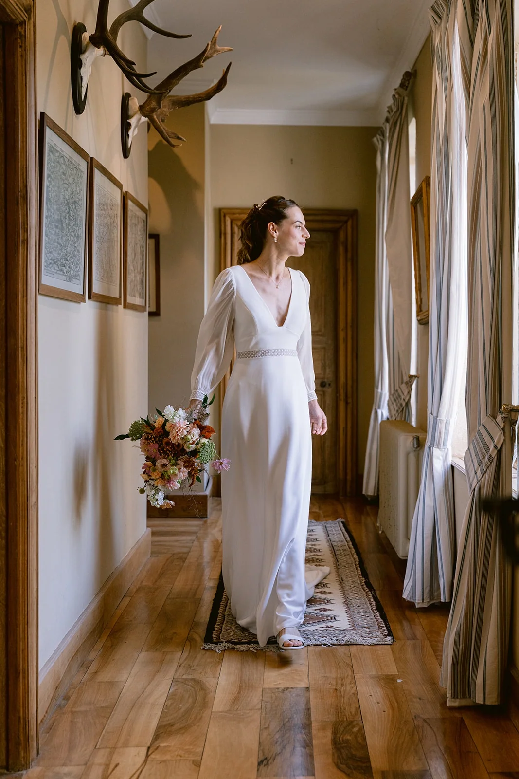 La mariée tient son bouquet dans un couloir au parquet ancien, éclairé par de grandes fenêtres.