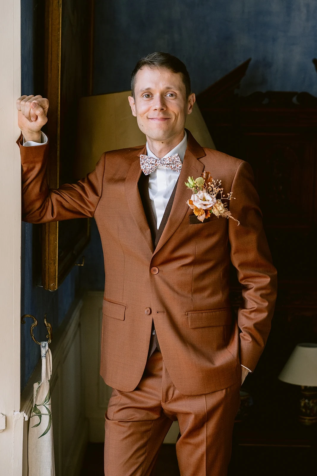 Le marié en costume brun, nœud papillon fleuri et boutonnière pose dans l’encadrement d’une porte.