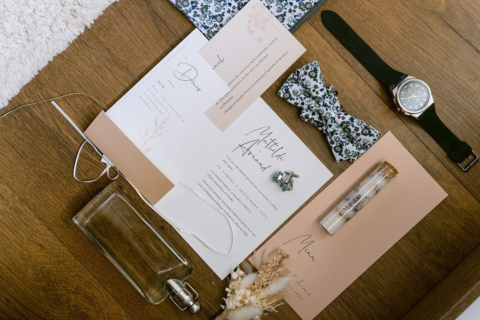 La papeterie, le nœud papillon fleuri, la montre et le parfum du marié sont posés sur une table.