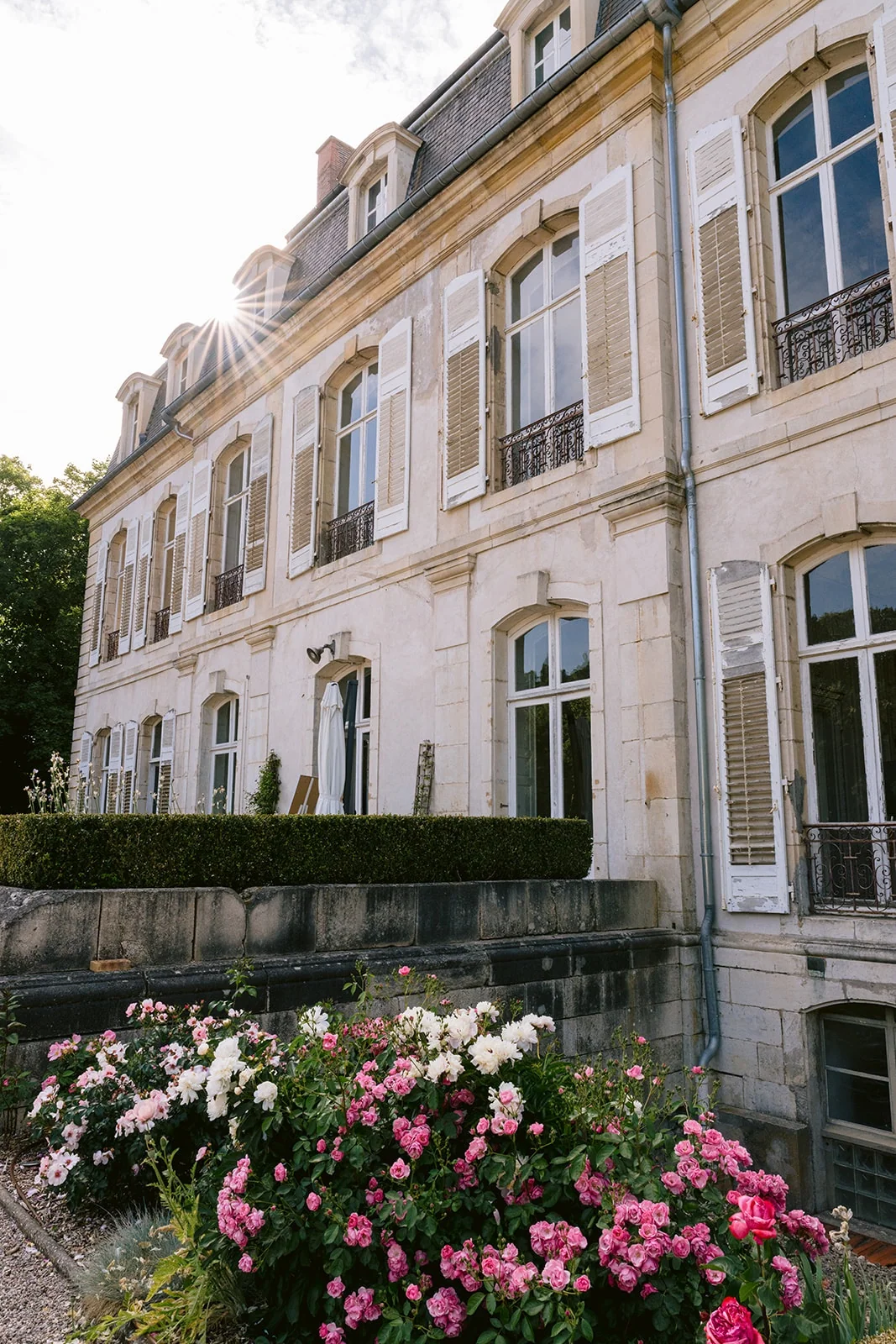 La façade en pierre d’une grande demeure, aux fenêtres et volets blancs, borde des massifs de roses.