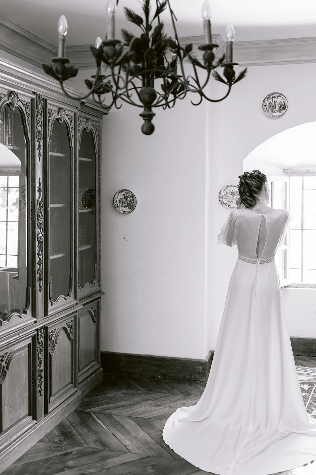 En noir et blanc, la mariée est vue de dos devant une fenêtre, sa longue traîne déployée sur le parquet.