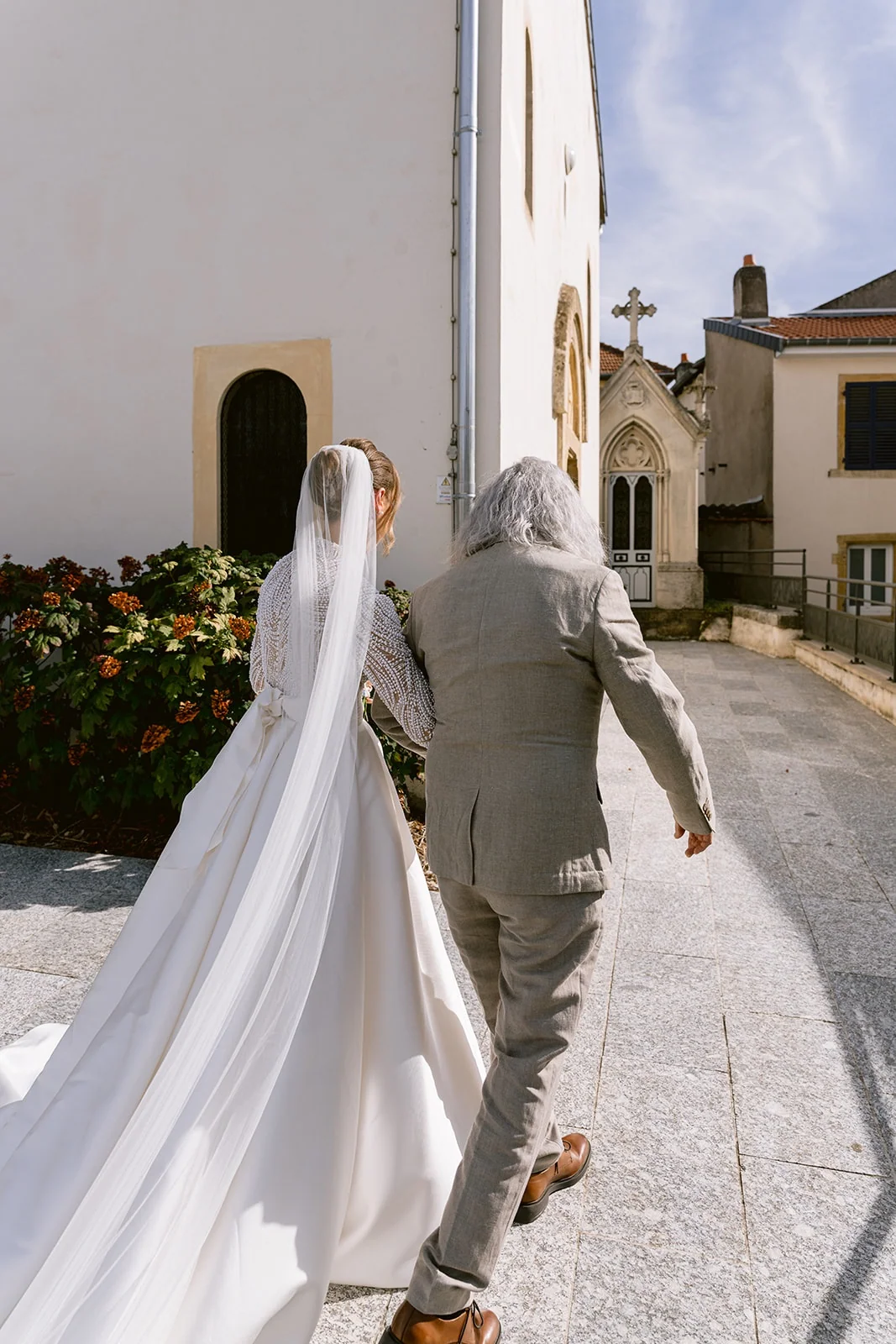 La mariée marche vers l’église au bras d’un proche, sa longue traîne derrière elle.