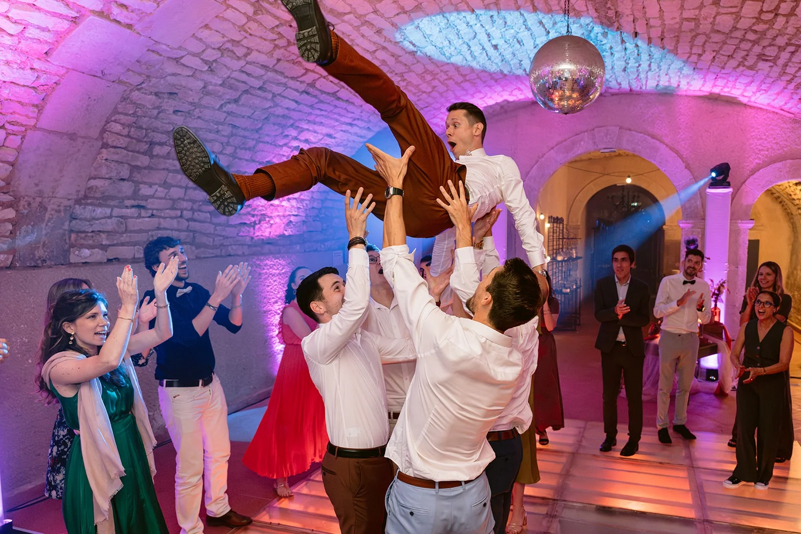 Des invités soulèvent le marié au-dessus de la piste de danse éclairée en rose et violet.
