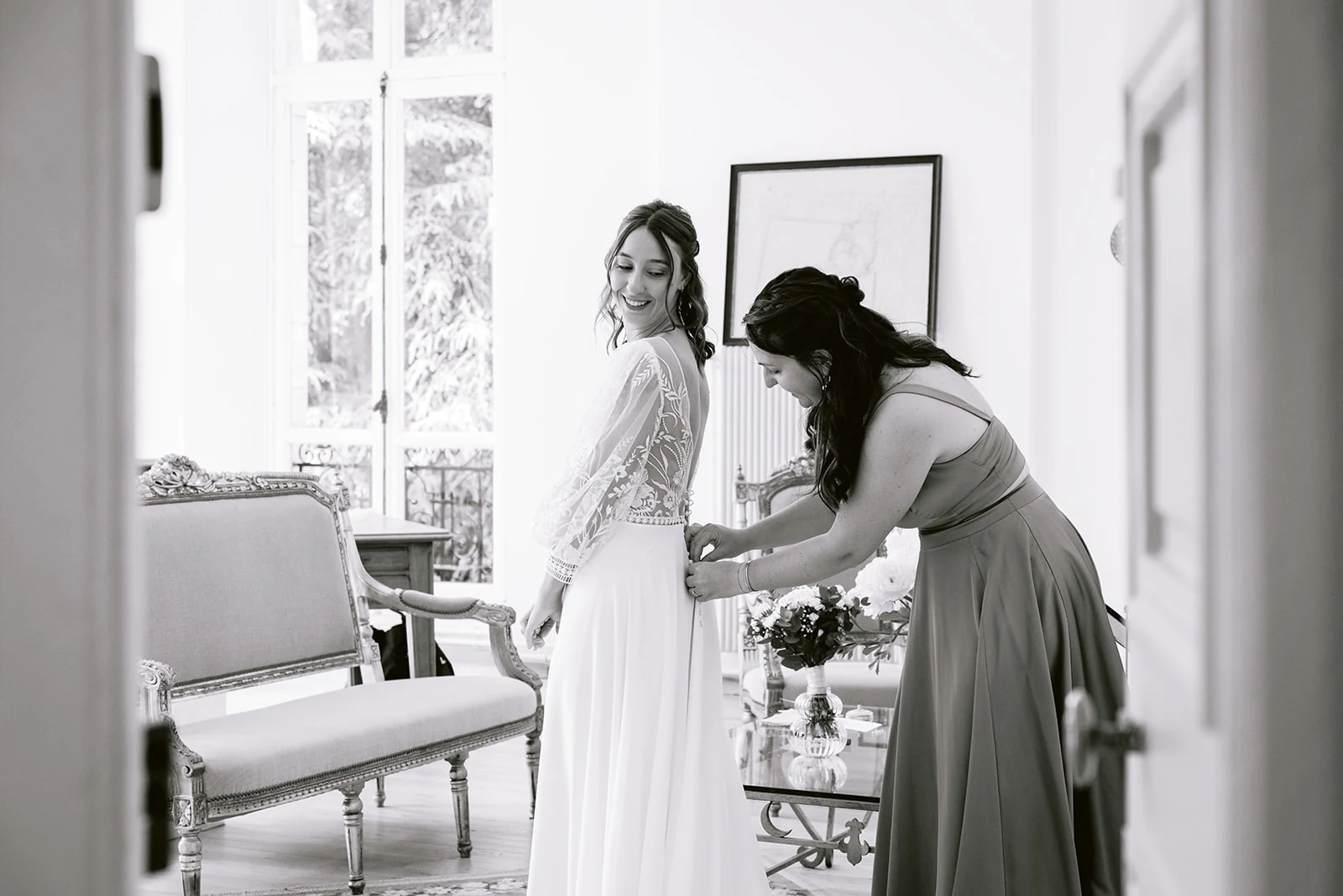 En noir et blanc, une femme ajuste le dos de la robe de la mariée, qui regarde par-dessus son épaule.