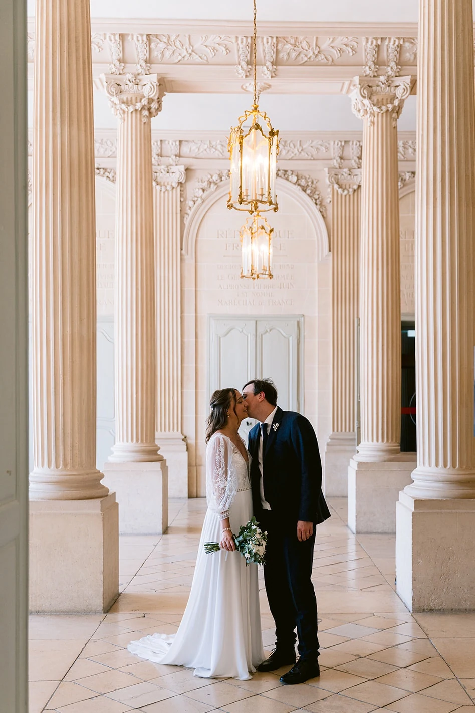 Les mariés s’embrassent dans un vaste hall aux colonnes blanches, sous des lustres dorés.