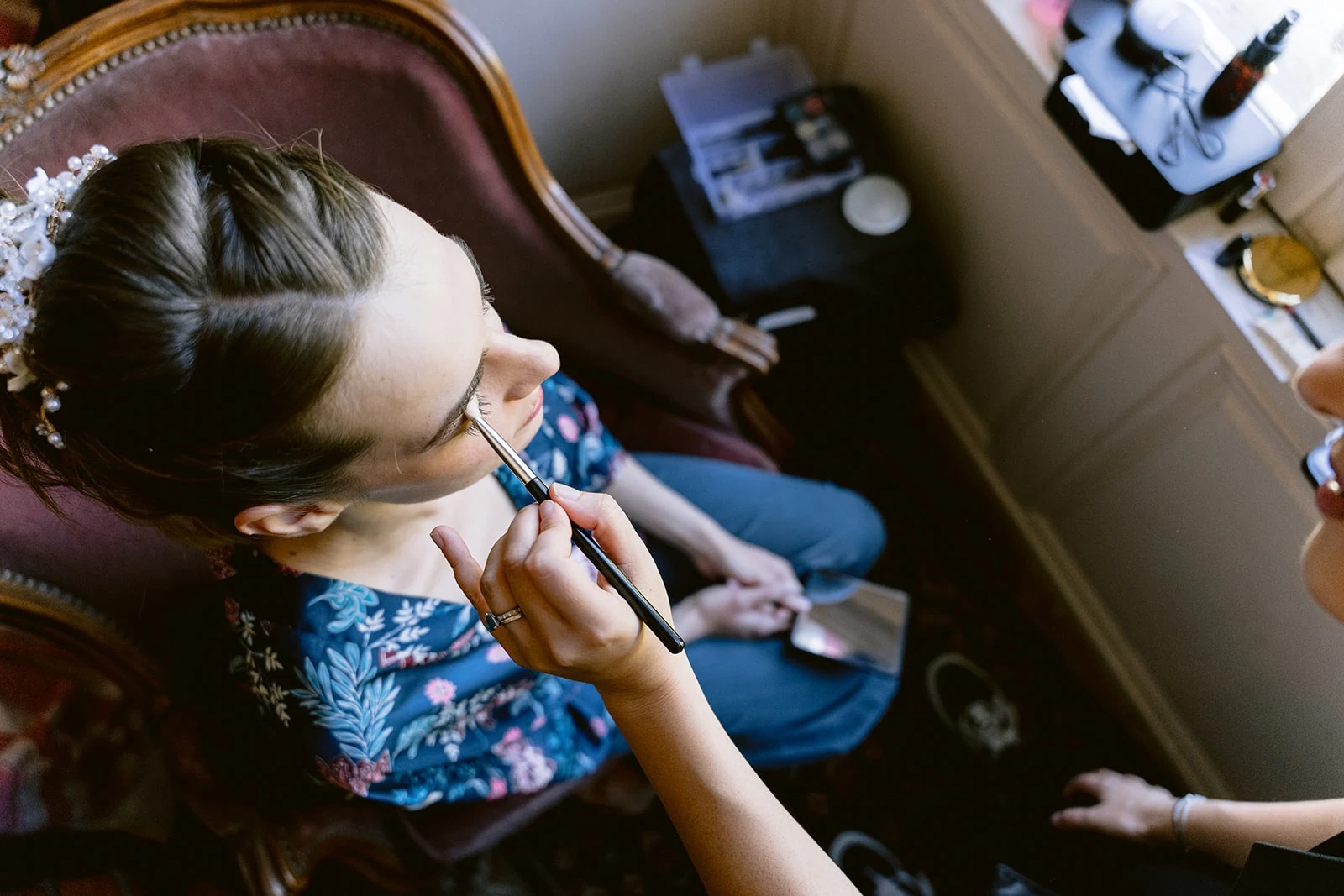 Vue de dessus, la mariée en peignoir fleuri se fait maquiller les yeux, assise dans un fauteuil.