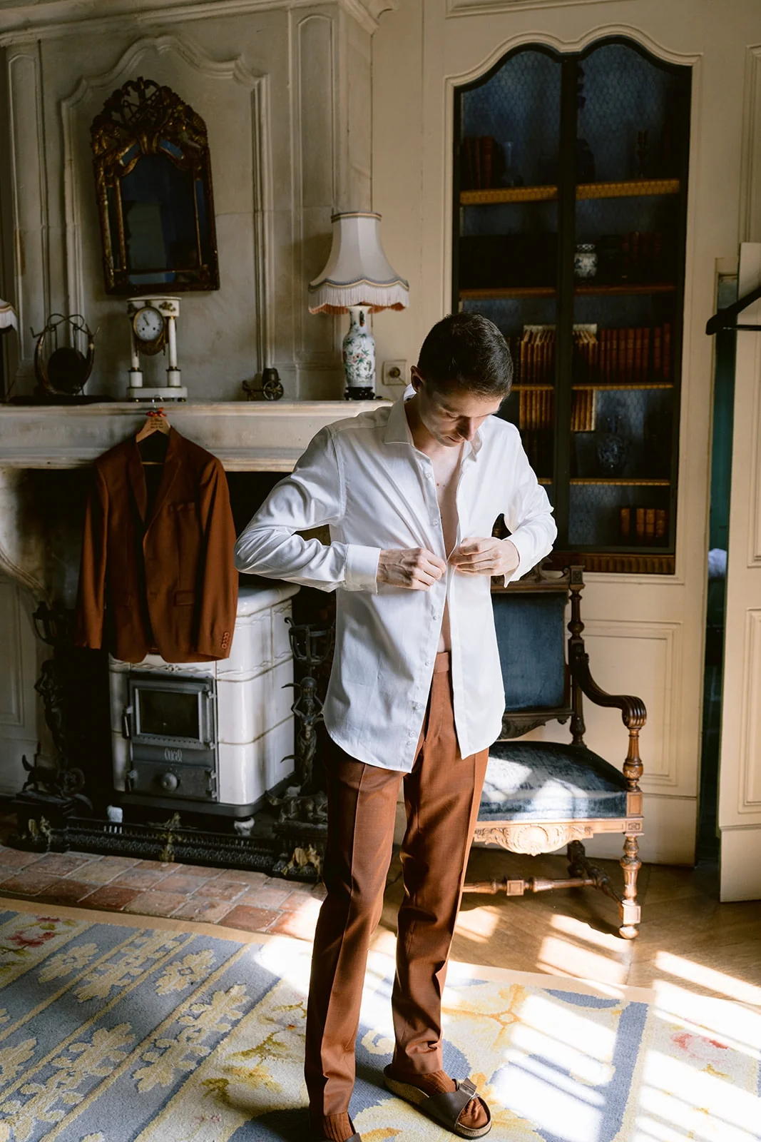 Le marié boutonne sa chemise blanche, vêtu d’un pantalon brun, dans une pièce éclairée par le soleil.