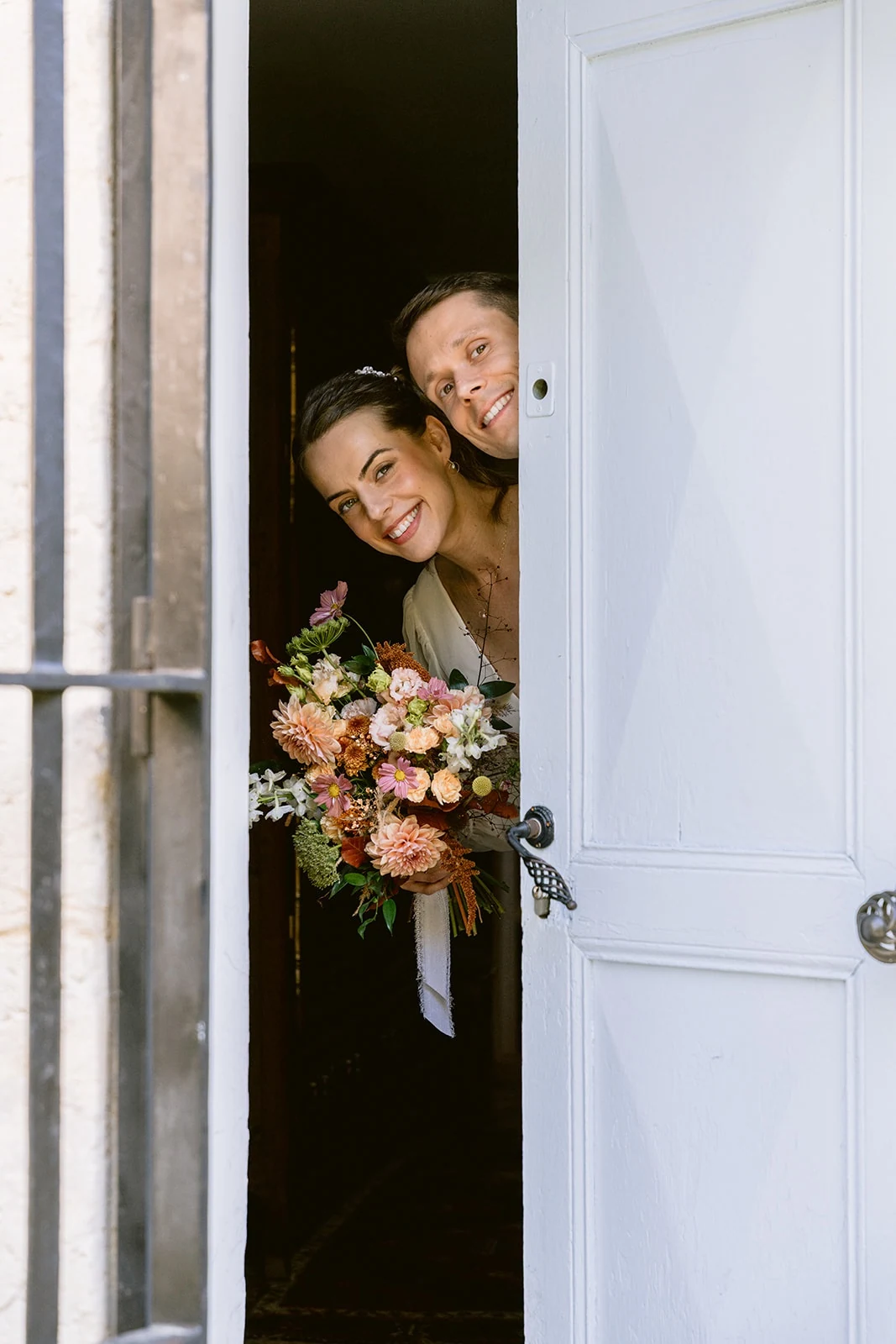 La mariée et une femme sourient en passant la tête par une porte entrouverte, un bouquet entre les mains.