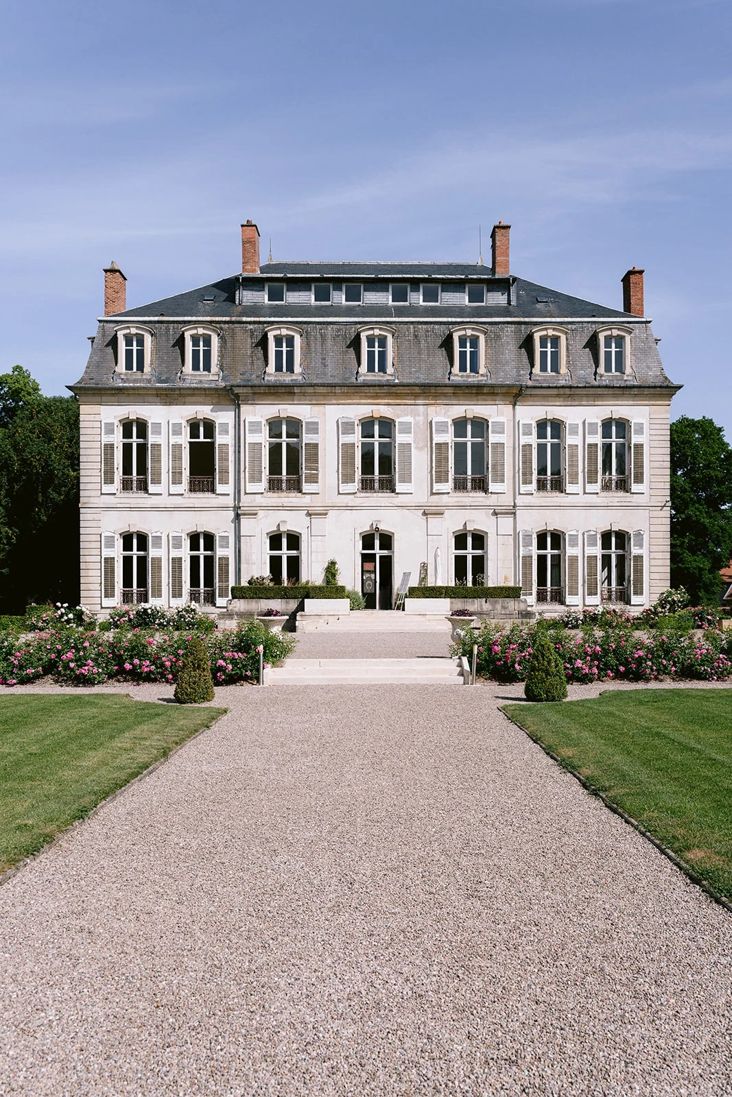 Une allée de gravier bordée de pelouses conduit à une grande demeure aux volets blancs et au toit mansardé.