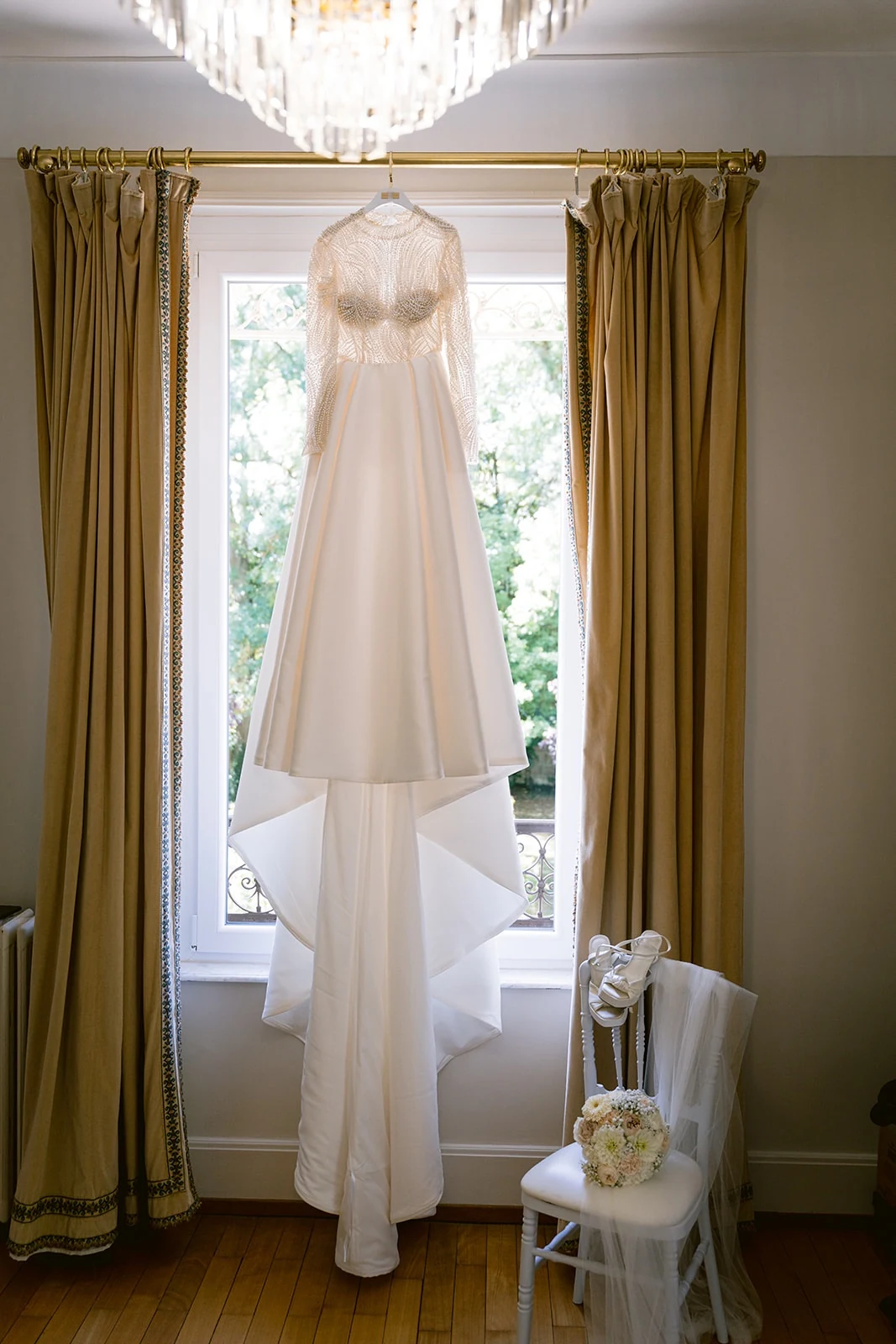 La robe de mariée est suspendue devant une fenêtre aux rideaux dorés.