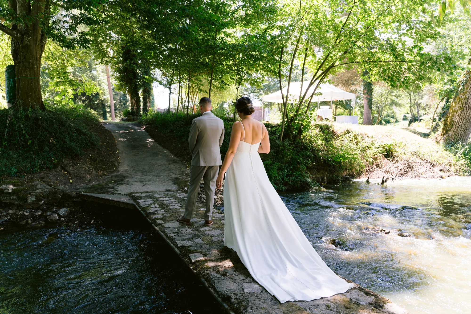 Les mariés avancent sur un passage au bord de l’eau, sous les arbres.