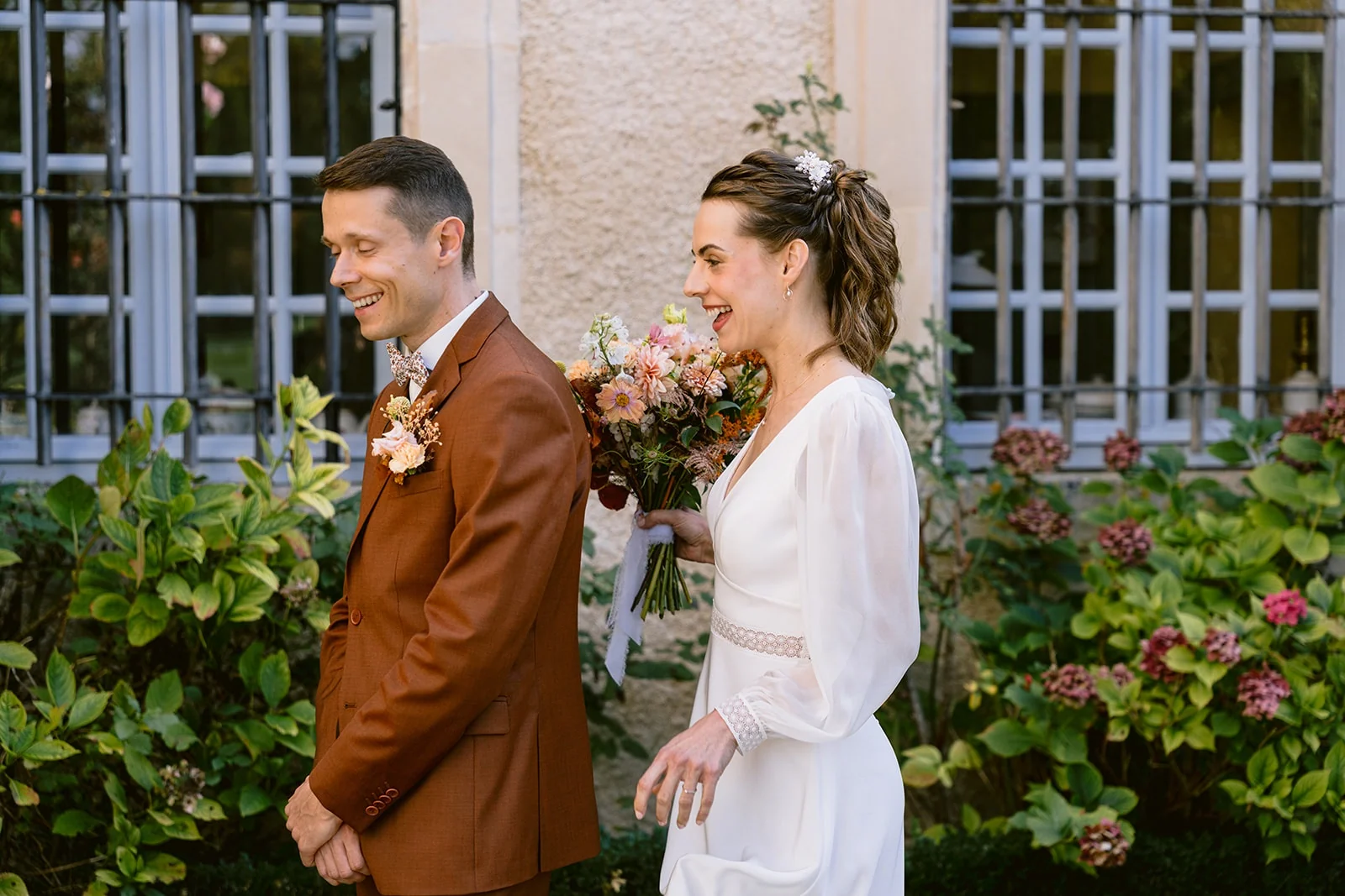 Le marié sourit tandis que la mariée se tient derrière lui avec son bouquet, devant une fenêtre fleurie.