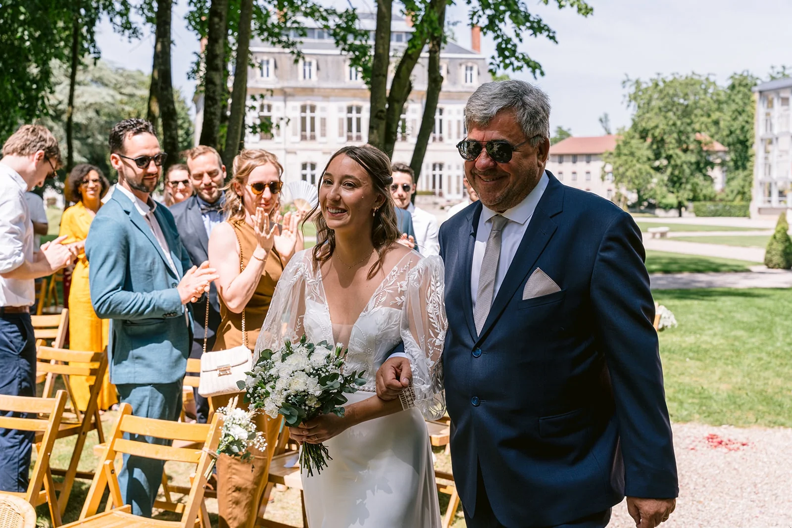 La mariée, bouquet à la main, avance au bras d’un homme en costume bleu entre les invités debout dans le jardin.