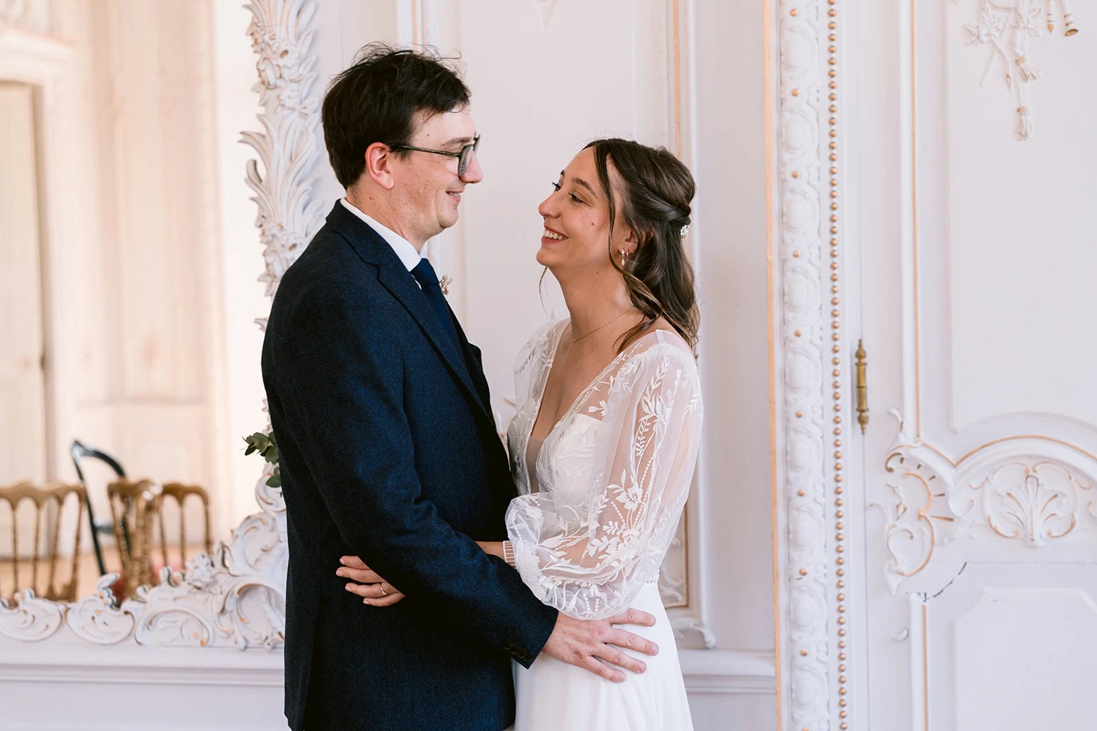 Les mariés se regardent en souriant, enlacés devant une porte blanche aux moulures décoratives.