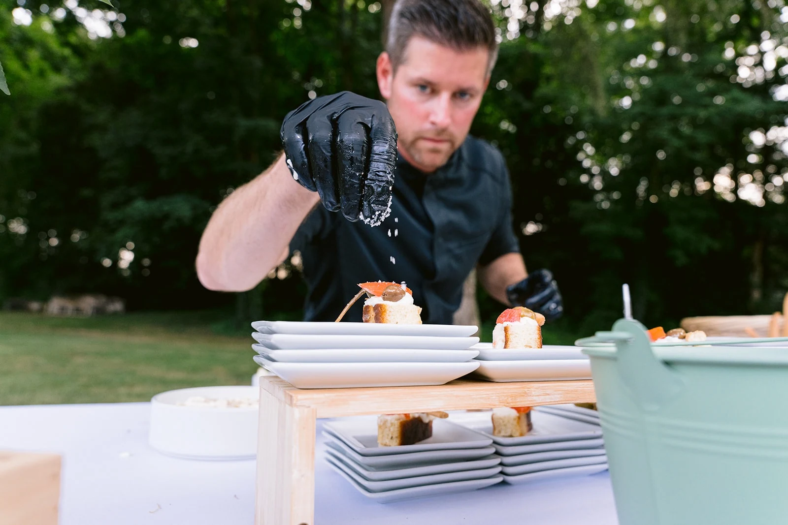 Un homme en tenue noire et gants termine le dressage de bouchées sur des assiettes, derrière un buffet extérieur.