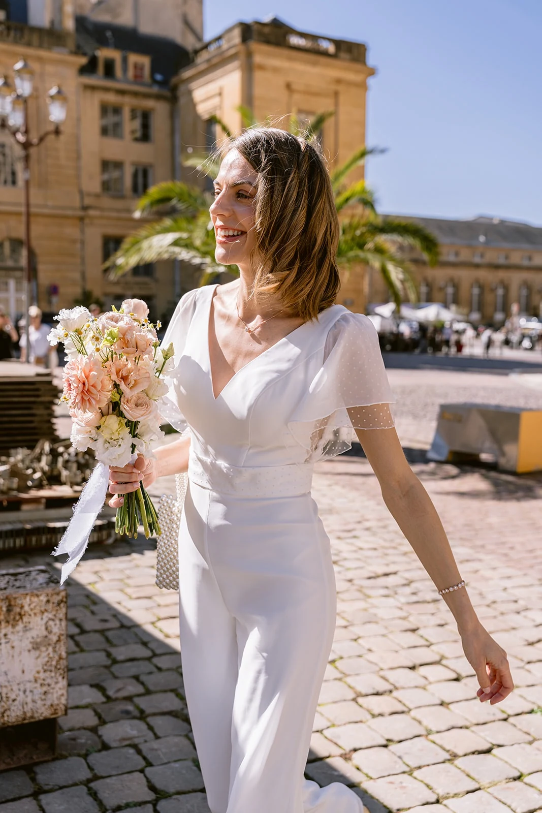 Une mariée en combinaison blanche sourit en tenant un bouquet rose et blanc sur une place pavée au soleil.