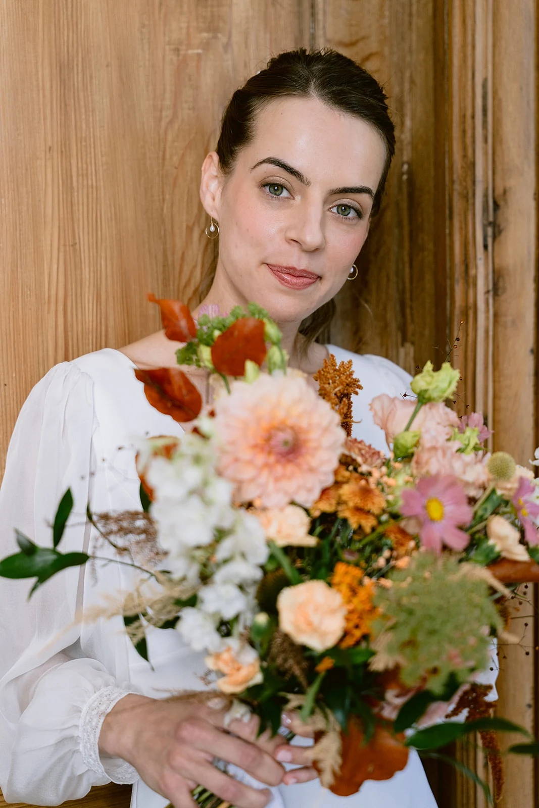 Portrait de la mariée devant une porte en bois, son bouquet de fleurs roses et orangées au premier plan.