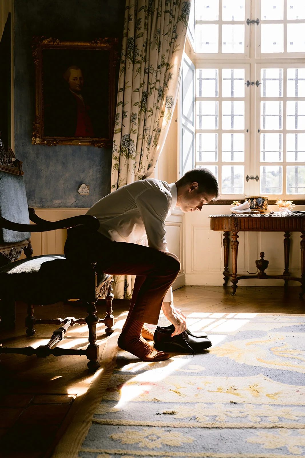 Le marié se penche pour mettre ses chaussures, assis près d’une grande fenêtre ensoleillée.