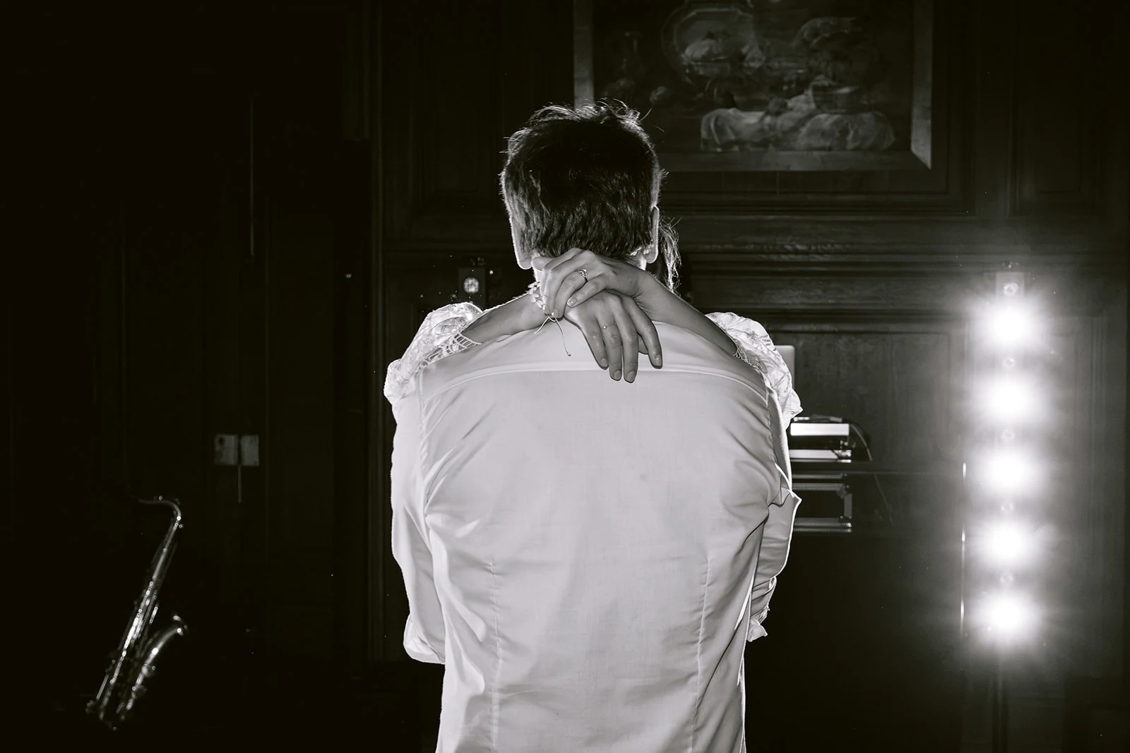 En noir et blanc, la mariée enlace le cou du marié, vu de dos en chemise blanche devant les lumières de la piste.