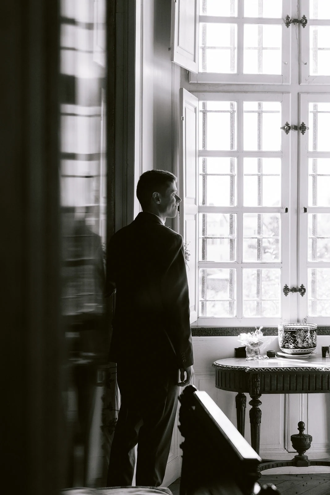 En noir et blanc, la silhouette du marié se détache devant une grande fenêtre lumineuse.