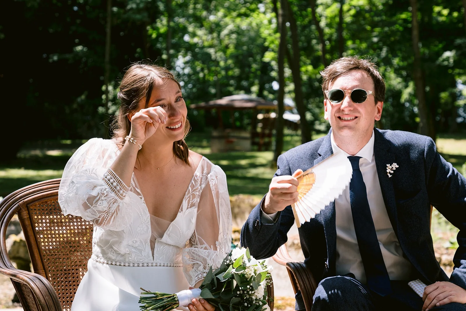 La mariée porte une main à son œil tandis que le marié sourit à côté d’elle, assis sous les arbres.