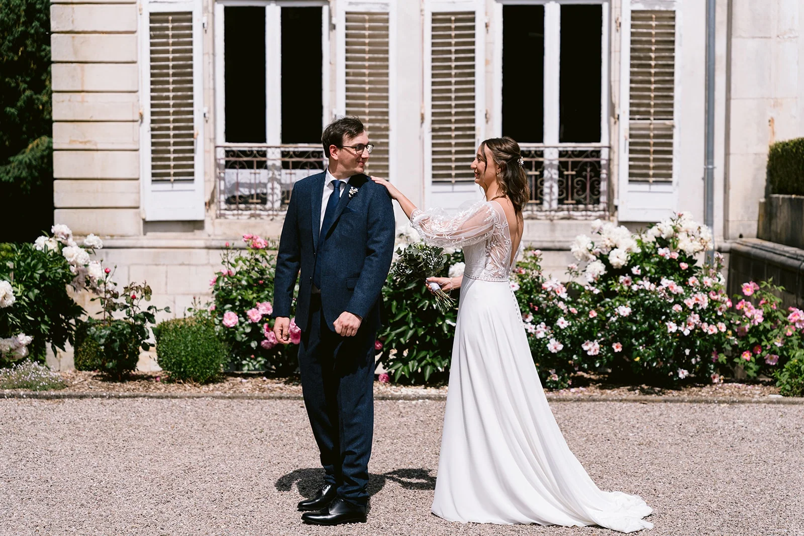 La mariée pose une main sur l’épaule du marié, qui se retourne vers elle dans la cour fleurie d’une demeure.