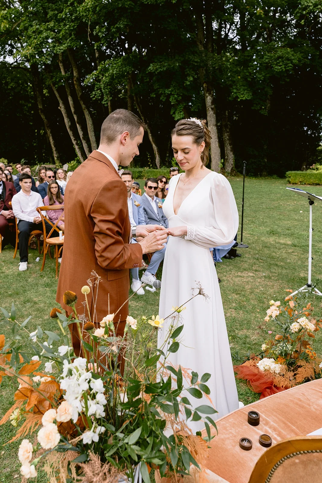 Le marié passe une alliance au doigt de la mariée, devant les fleurs de la cérémonie en plein air.