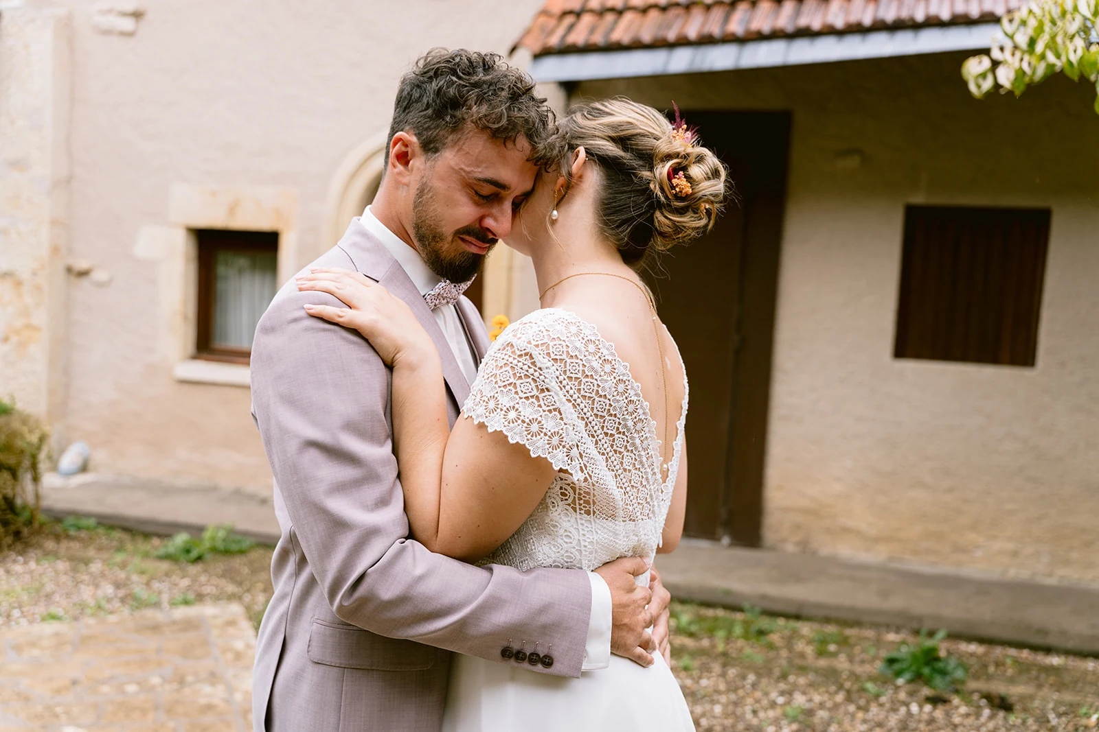 Le marié enlace la mariée tandis qu’elle se penche à son oreille, dans une cour.