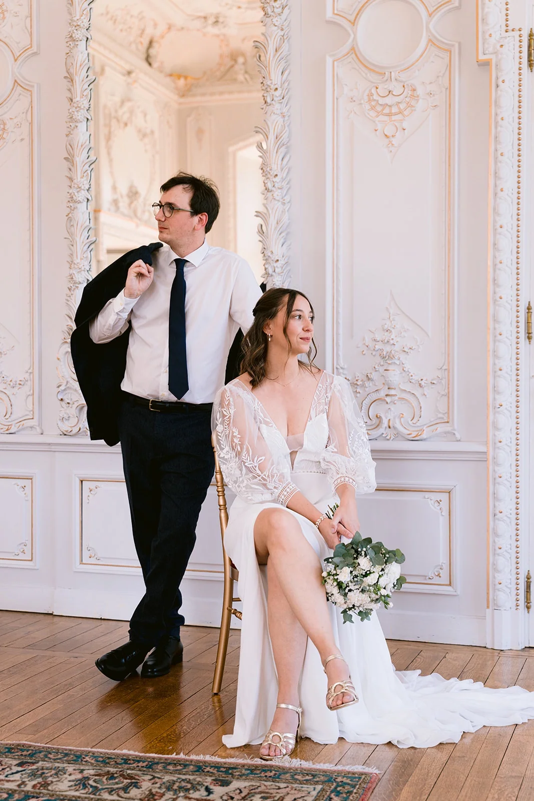 La mariée est assise avec son bouquet, près du marié debout, dans un salon aux moulures blanches et dorées.