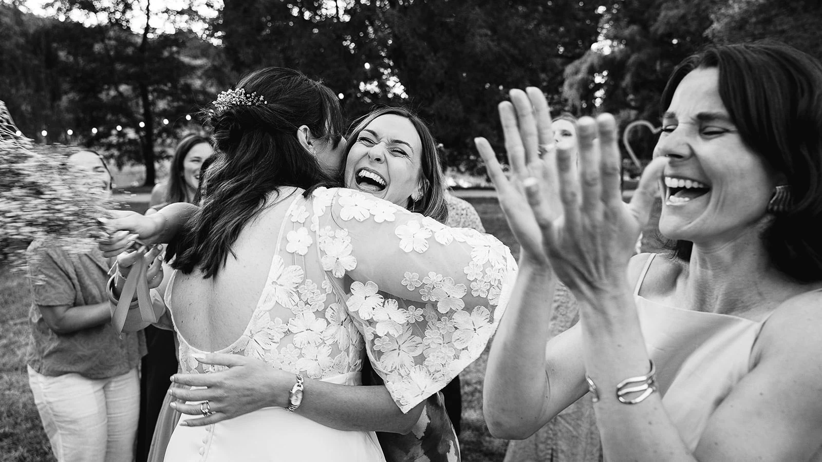 En noir et blanc, la mariée serre une proche dans ses bras, entourée de rires.