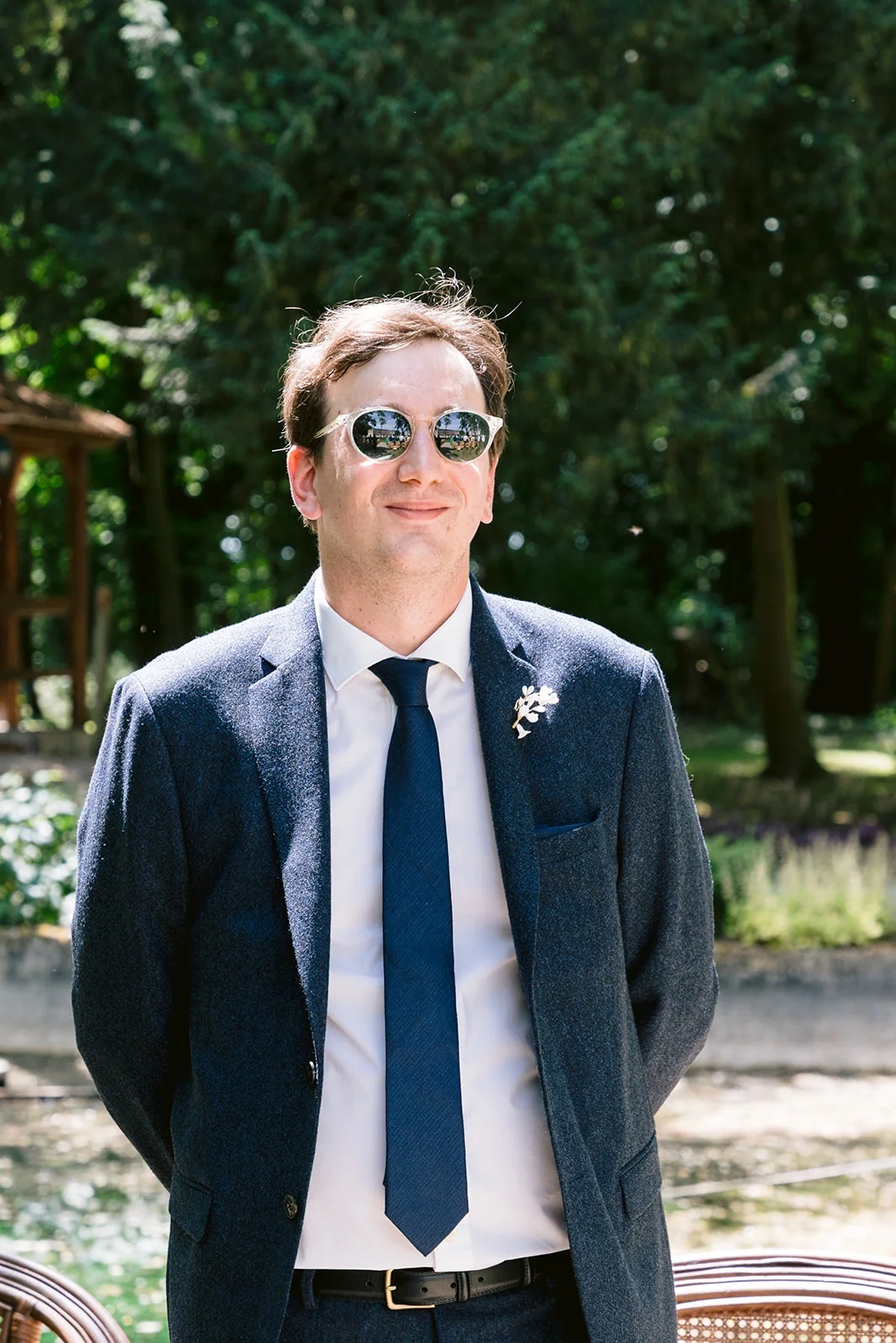 Le marié en costume bleu et lunettes de soleil se tient debout sous les arbres, les mains derrière le dos.