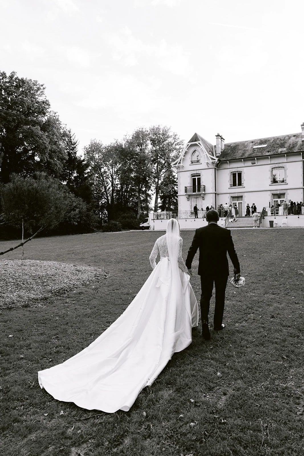 En noir et blanc, les mariés marchent vers la demeure, vus de dos.