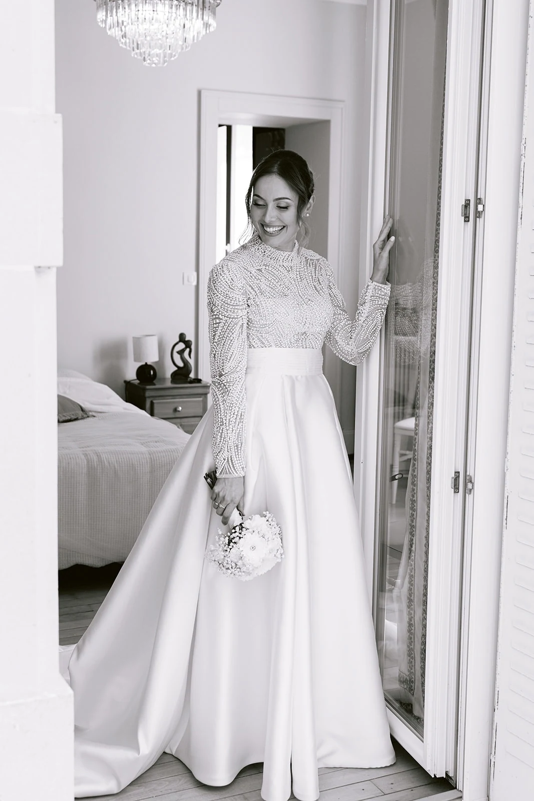 En noir et blanc, la mariée baisse les yeux vers son bouquet près d’une porte ouverte.