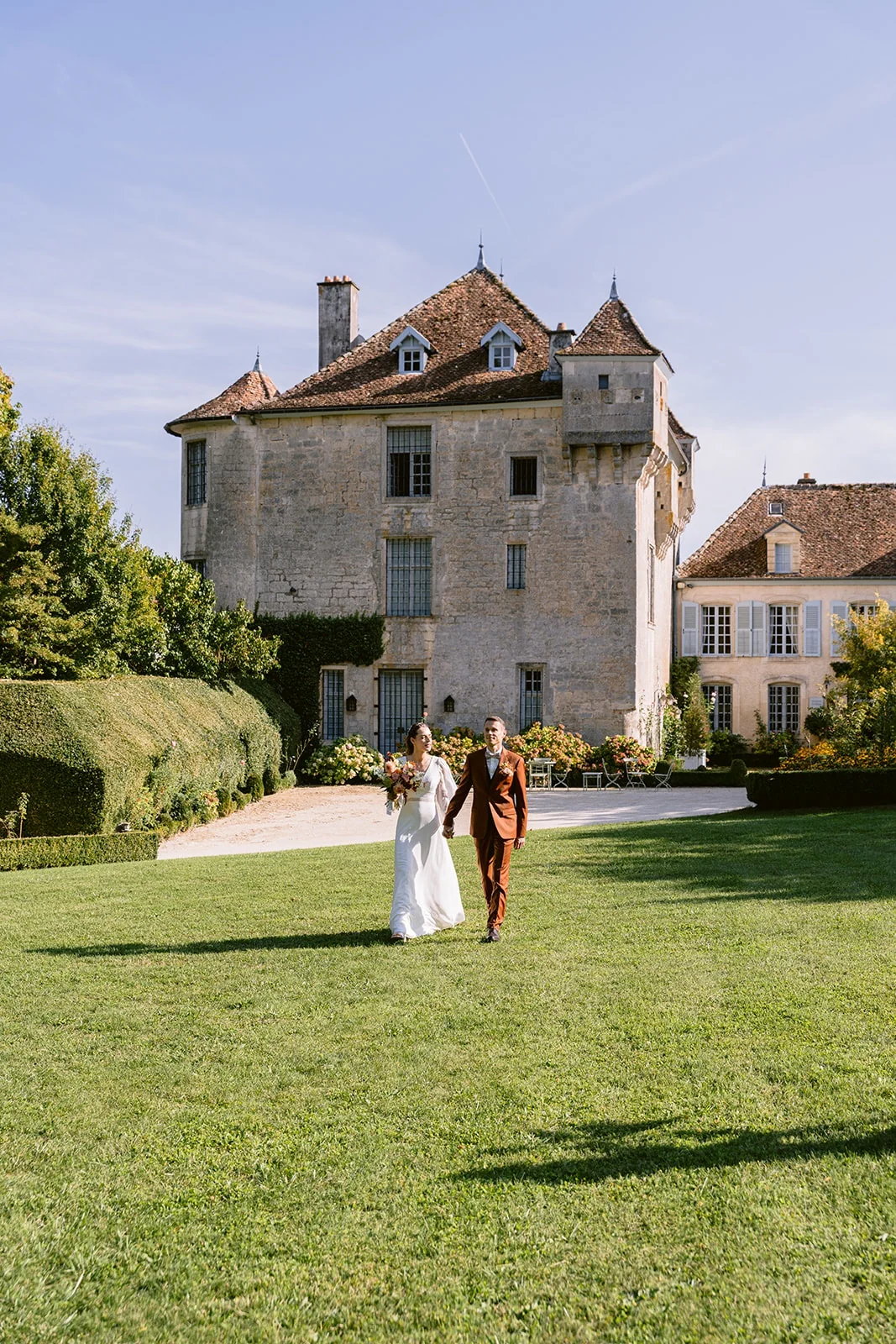 Les mariés avancent sur une grande pelouse devant une demeure en pierre aux toits pointus.