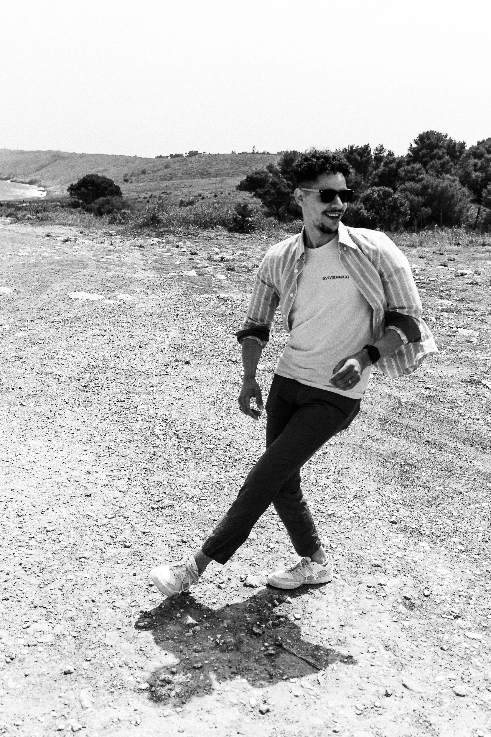 Portrait en noir et blanc d’un homme en lunettes de soleil, marchant sur un sentier caillouteux.
