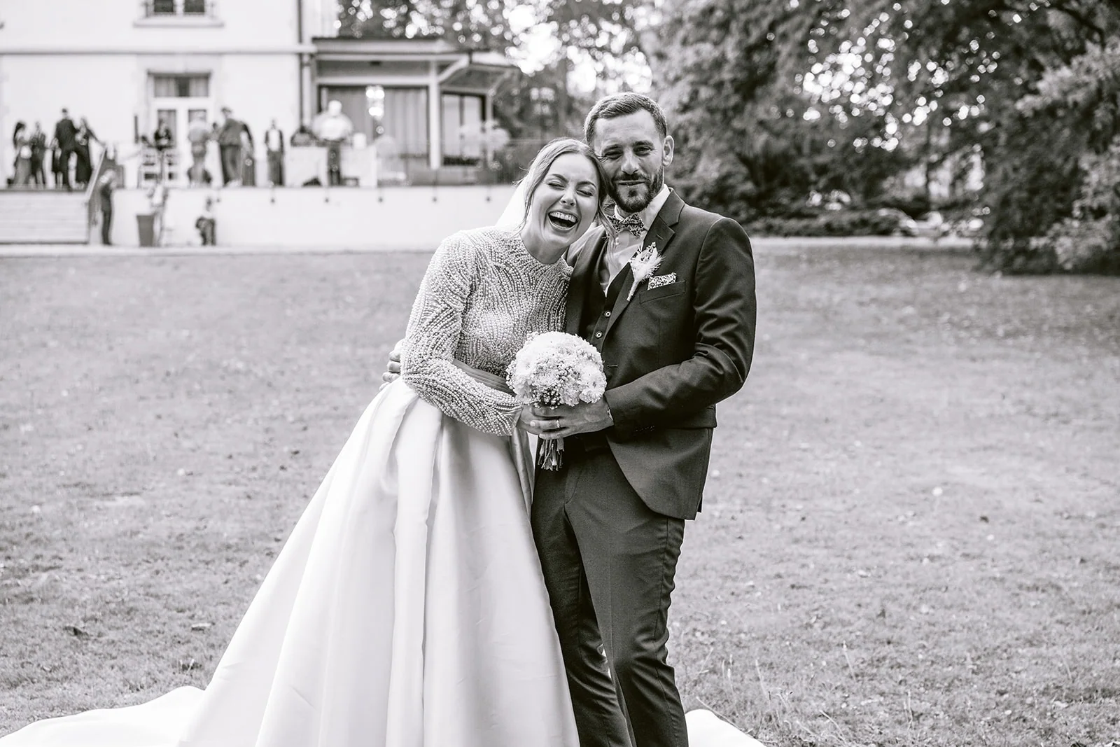 En noir et blanc, les mariés éclatent de rire devant la demeure.