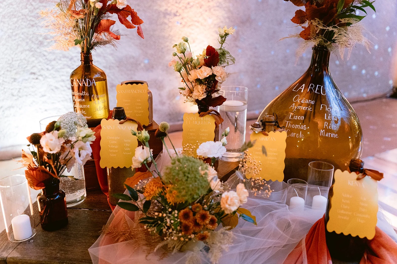 Des bouteilles ambrées, des fleurs et des cartons orangés décorent une table recouverte de voilages.