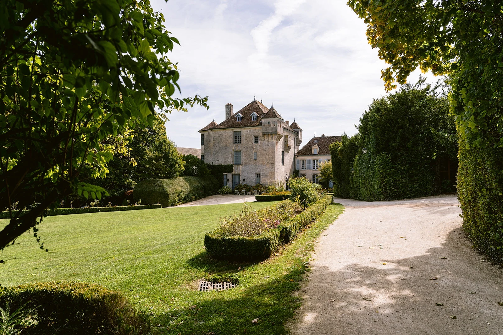 Un château de pierre au bout d’une allée de gravier bordée de pelouse, de haies et d’arbres.