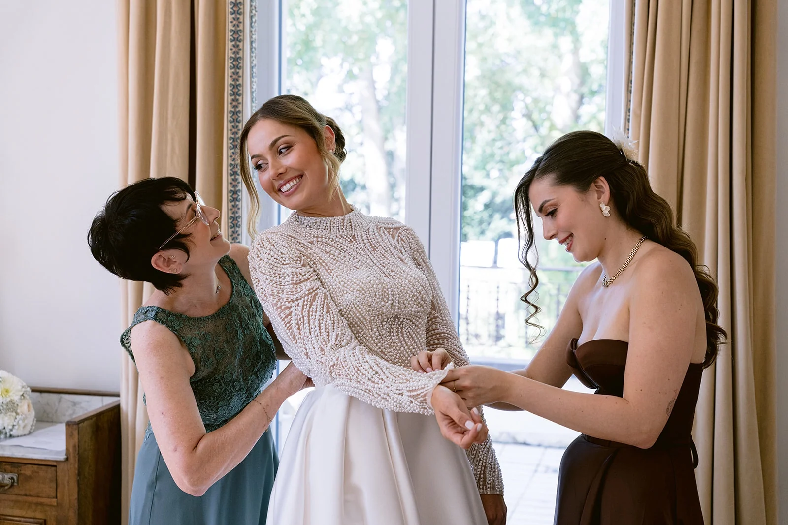 Deux proches aident la mariée à enfiler sa robe à manches en dentelle.