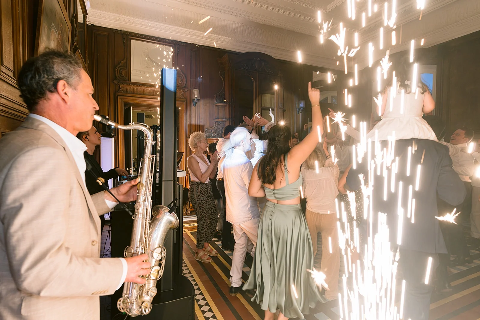 Un saxophoniste joue près des invités qui dansent, au milieu de jets d’étincelles lumineux.