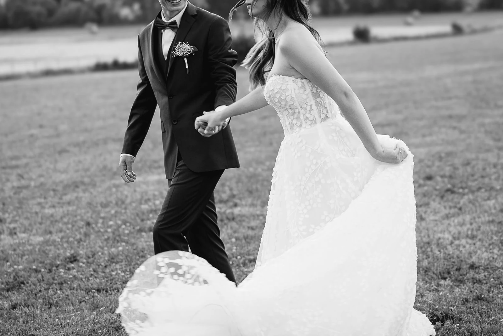 En noir et blanc, les mariés marchent dans l’herbe, la mariée soulevant sa jupe de dentelle.