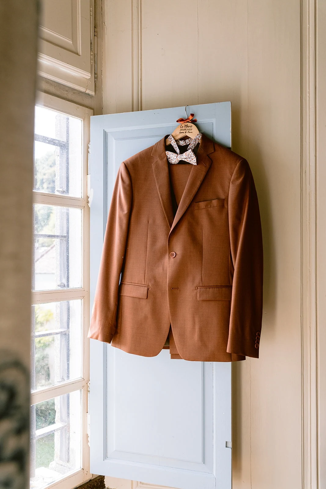 Une veste de costume brune et un nœud papillon sont suspendus devant une fenêtre ouverte.