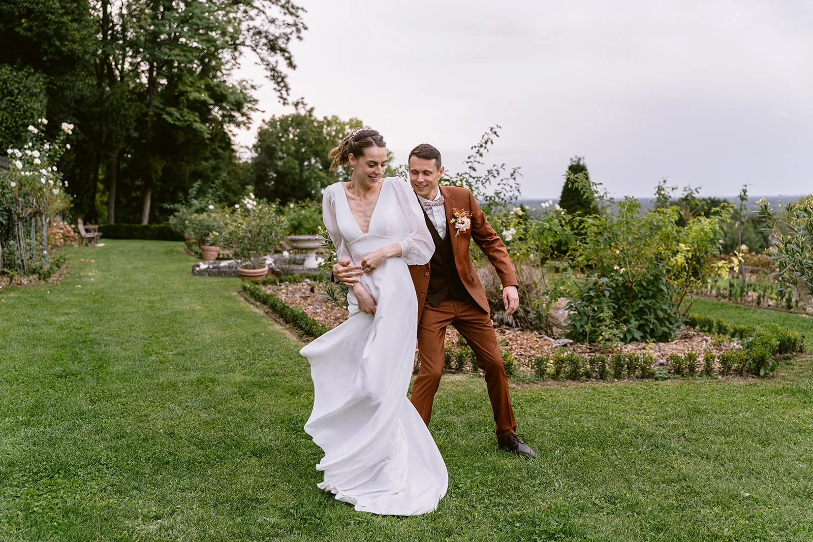 La mariée soulève le bas de sa robe en avançant sur la pelouse, suivie du marié souriant.