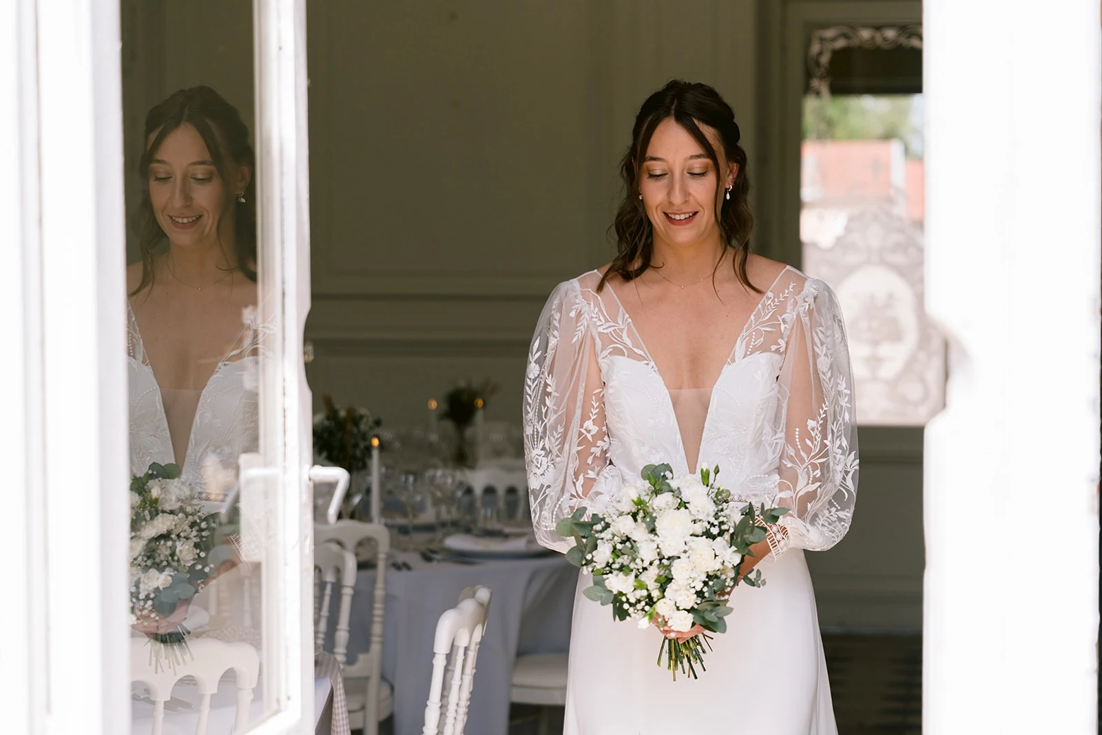 La mariée tient son bouquet blanc dans l’encadrement d’une porte vitrée, où se reflète sa silhouette.