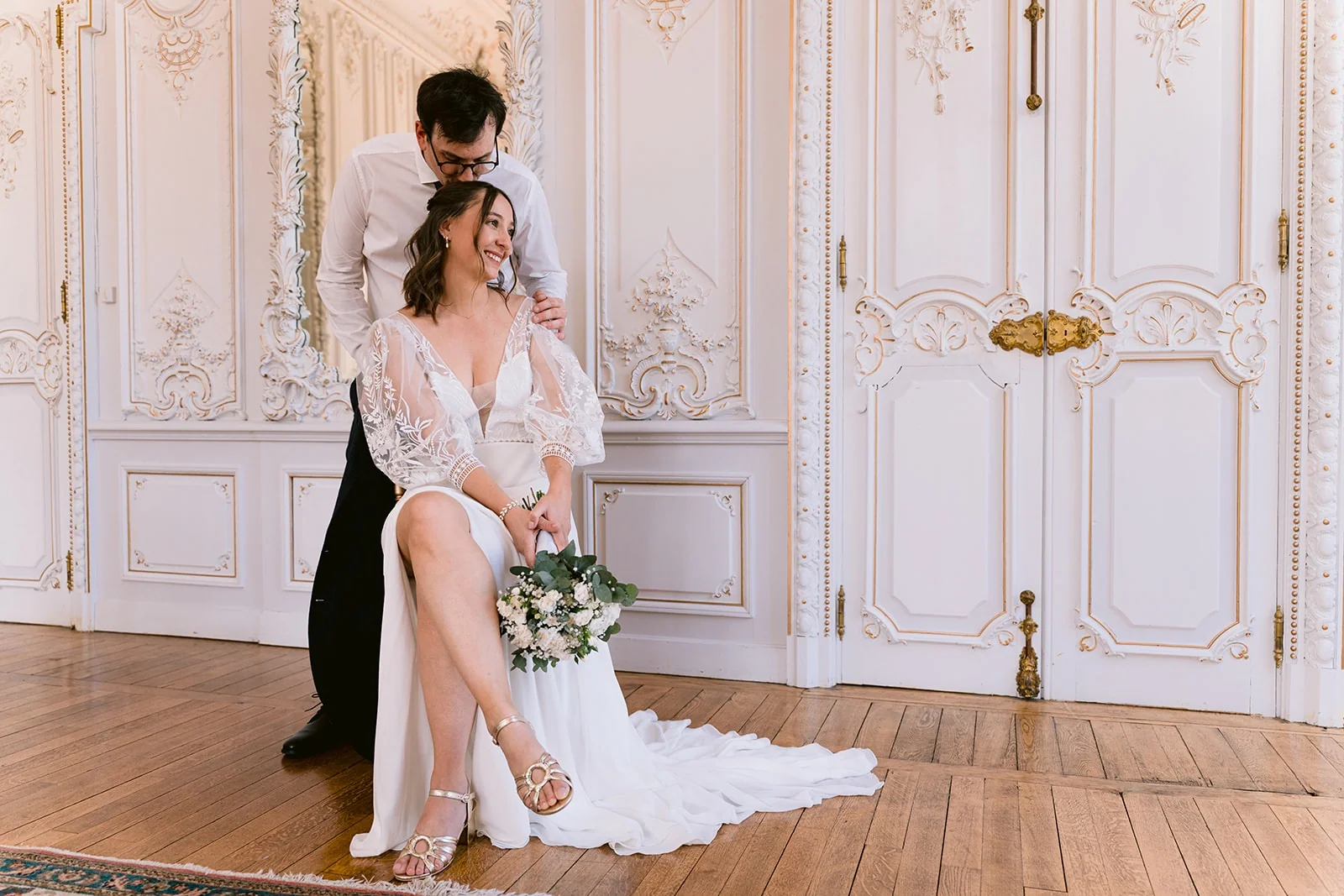 Le marié embrasse la tête de la mariée, assise avec son bouquet devant des boiseries blanches.