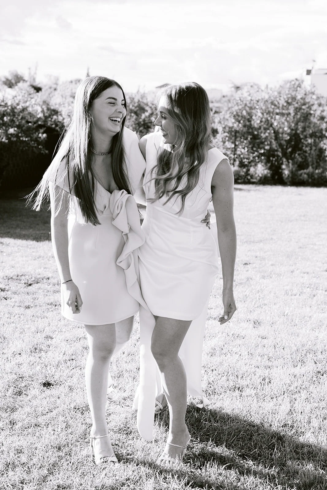 En noir et blanc, deux femmes en robe blanche rient ensemble dans le jardin.