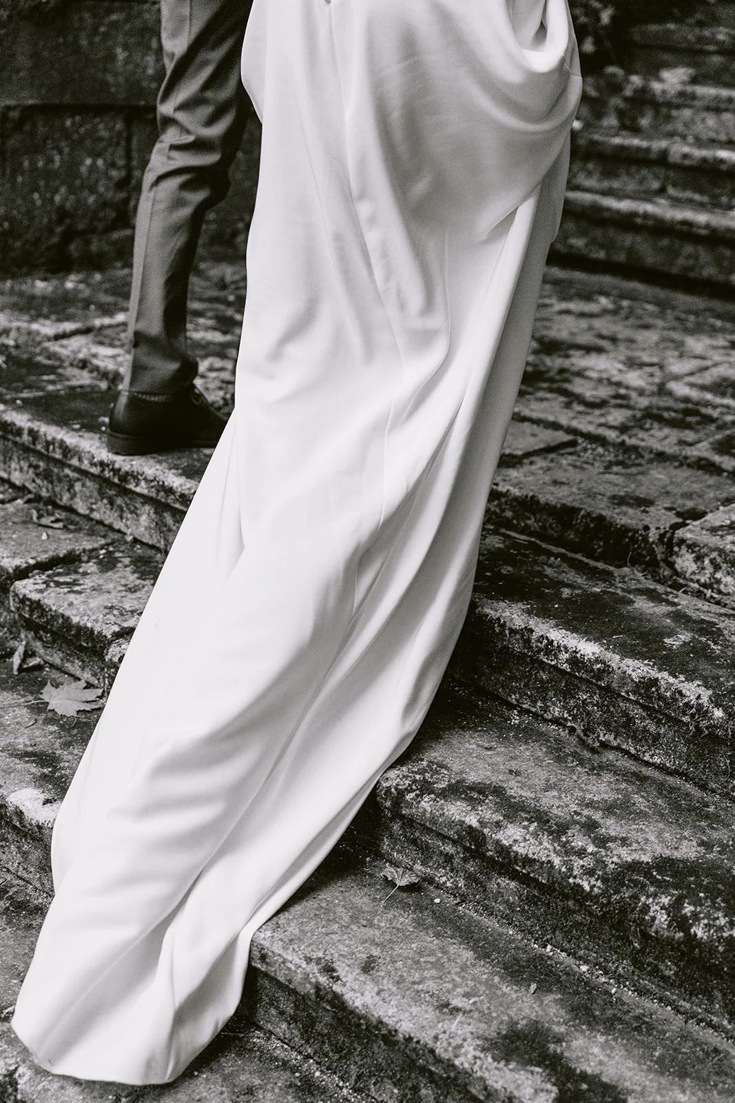 En noir et blanc, la traîne de la robe de mariée glisse sur les marches d’un escalier en pierre.