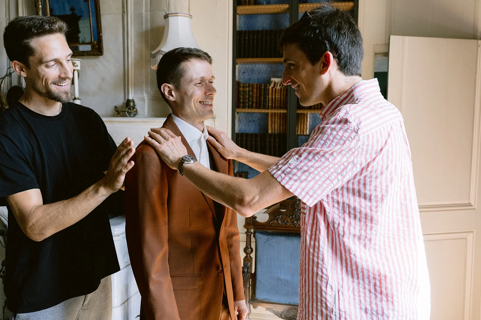 Deux hommes aident le marié souriant à enfiler sa veste de costume brune.